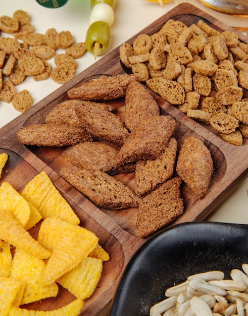 Free photo side view of various salty beer snacks bread crackers corn cones on a wood platter and sunflowers seeds and pickled olives on white