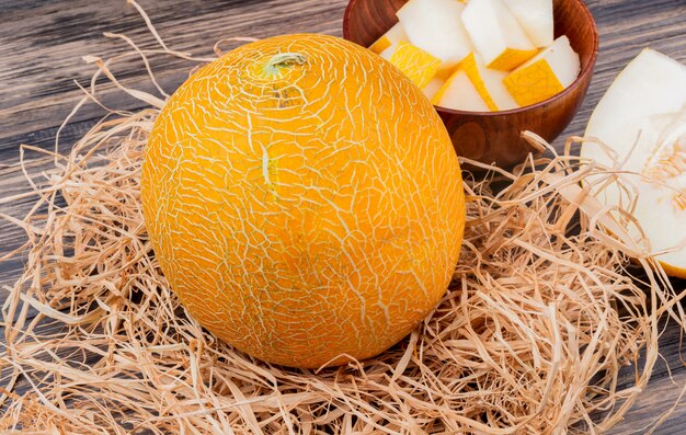 Side view of whole melon on straw with melon slices in bowl and cut one on wooden background