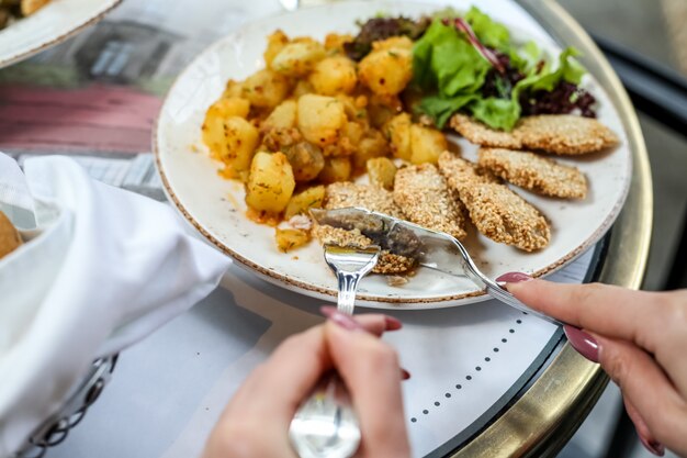 Side view woman eats chicken nuggets with potatoes and salad leaves on a plate