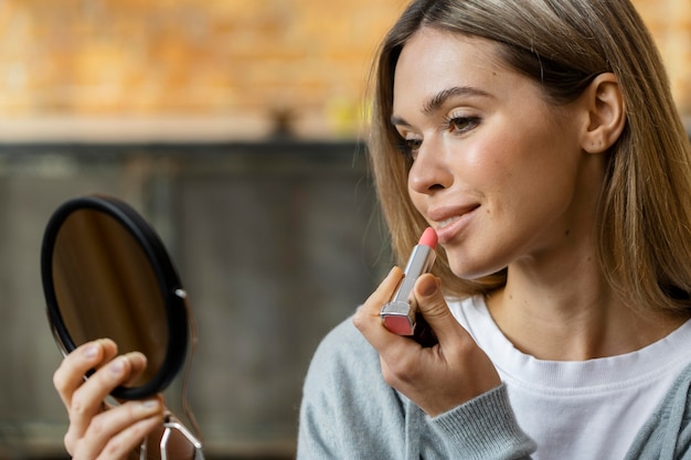 Free photo side view of woman looking in the mirror while putting on lipstick