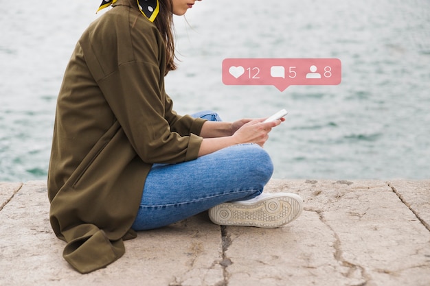 Free photo side view of woman sitting on pier using social media application on mobile phone