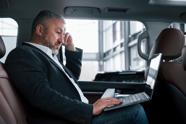 Free photo side view. working on a back of car using silver colored laptop. senior businessman