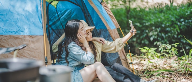 Free Photo side view young asian pretty woman and her girlfriend sitting at front of tent use mobile phone take photo during camping in forest with happiness together