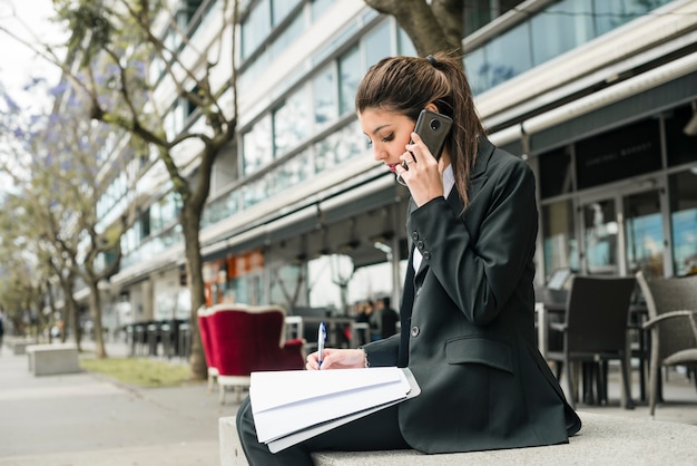 Free photo side view of a young businesswoman sitting outside the building writing on folder with pen