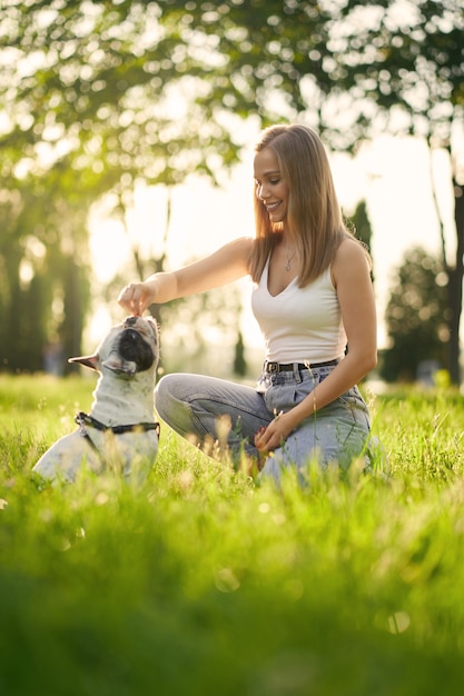 Side view of young smiling woman training french bulldog in city park. Purebred pet smelling treats from hand of female dog owner, beautiful summer sunset on background. Animal training concept.