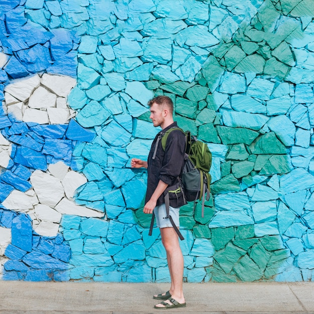 Free Photo side view of young traveler man carrying backpack against stone wall