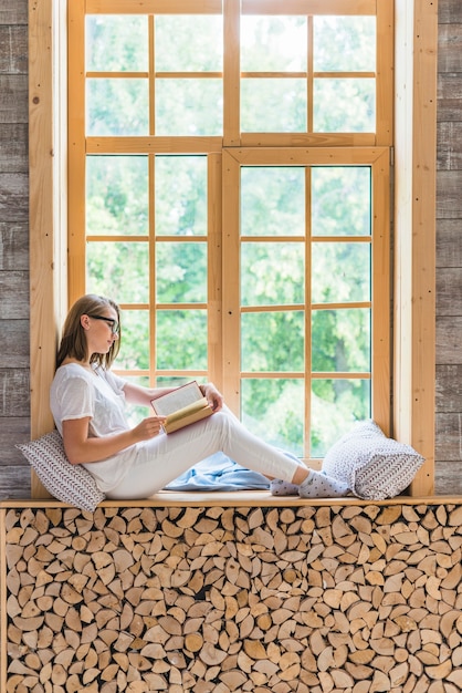 Free Photo side view of young woman reading book sitting near the window sill