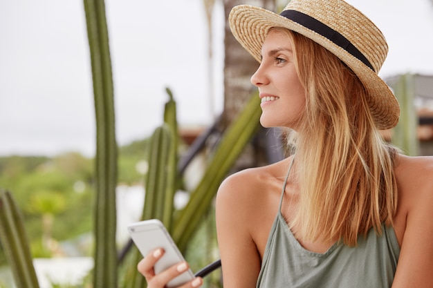 Sideways portrait of happy lovely woman with dreamy positive look, wears summer hat