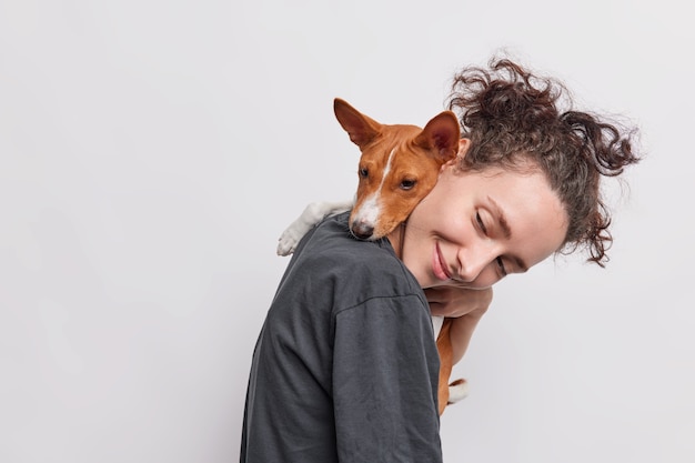 Sideways shot of adult girl carries puppy which embraces her around neck walkk together isolated over white 