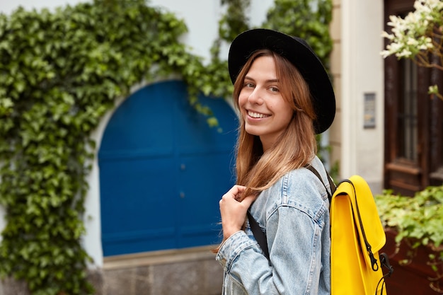 Sideways shot of cheerful lovely female strolls across old street with green vegetation, wears fashionable hat and denim jacket