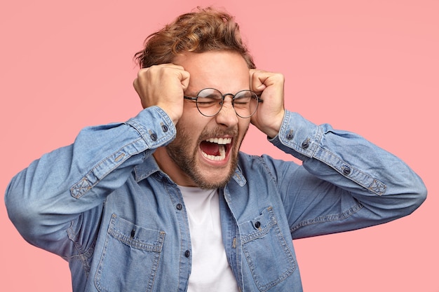 Free photo sideways shot of desprerate stressful guy keep fists on temples, screams angrily, frowns in discontent, expresses negative feelings