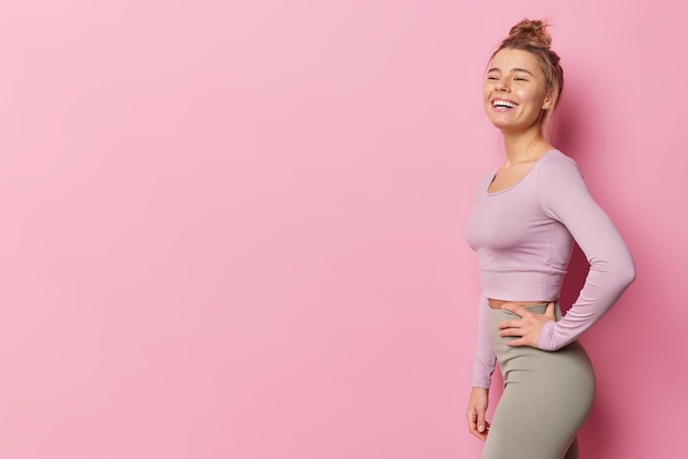 Free Photo sideways shot of healthy slim woman dressed in sportswear laughs happily has broad smile satisfied after exercising has regular fitness training poses against pink background blank copy space