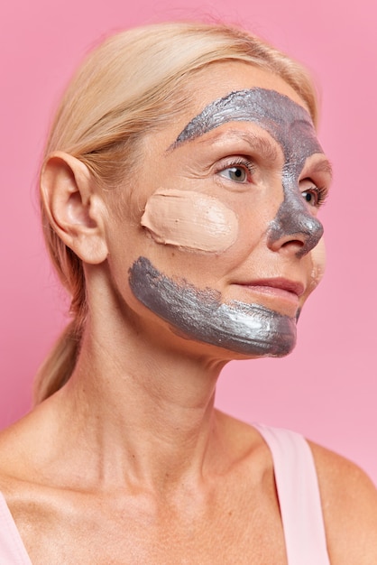 Sideways shot of serious European woman with combed fair hair undergoes cosmetic procedures to rejuvenate and nourish skin applies silver mask focused into distance poses against pink wall