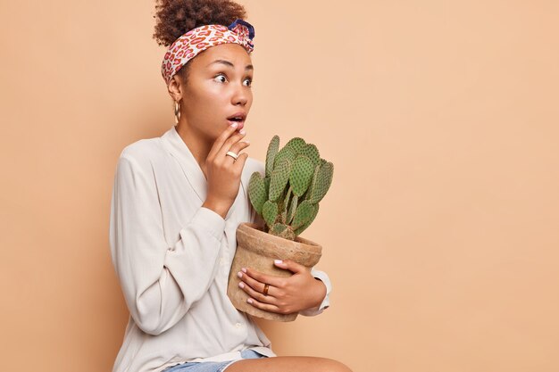 Sideways shot of surprised woman stares impressed holds breath holds potted cactus feels impressed wears kerchief tied over head white shirt isolated over beige wall copy space