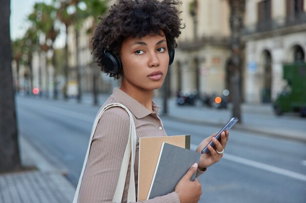 Sideways shot of thoughtful female student uses smartwatch and headphones listens educational webinar via headphones while goes to university holds notepads looks away with pensive expression