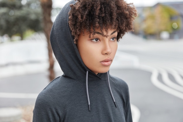 Sideways shot of thoughtful woman with dark skin, curly hair, wears hoodie