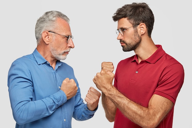 Free Photo sideways shot of two men competitors look seriously at each other, keeps hand clenched in fists, ready to fight, can`t share common business, stand against white wall. people and competition