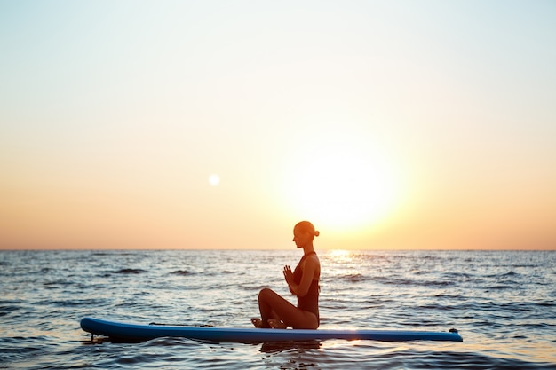 Silhouette of beautiful woman practicing yoga on surfboard at sunrise.