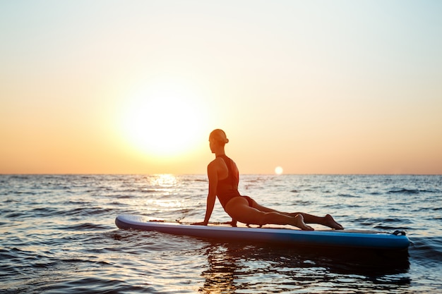 Free photo silhouette of beautiful woman practicing yoga on surfboard at sunrise.