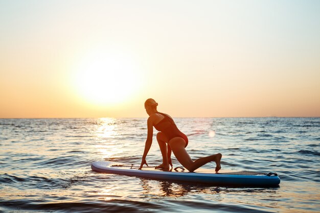 Silhouette of beautiful woman practicing yoga on surfboard at sunrise.