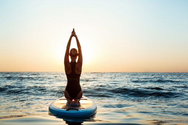 Silhouette of beautiful woman practicing yoga on surfboard at sunrise.