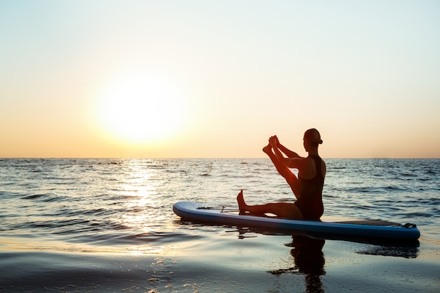 Silhouette of beautiful woman practicing yoga on surfboard at sunrise.