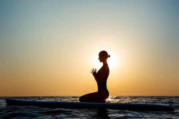 Free photo silhouette of beautiful woman practicing yoga on surfboard at sunrise.