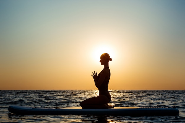 Silhouette of beautiful woman practicing yoga on surfboard at sunrise.