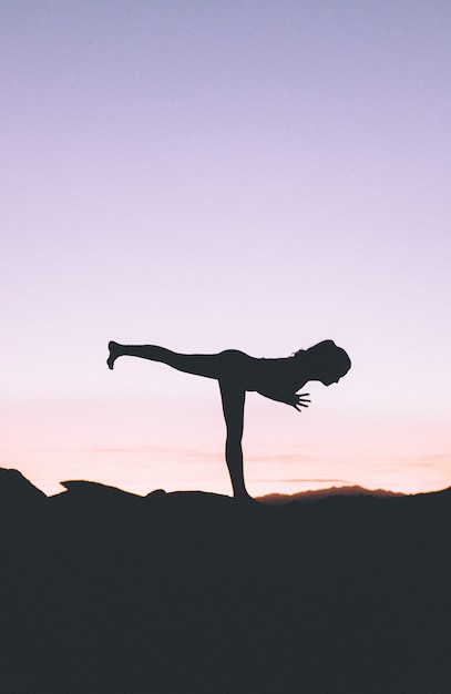 Free photo silhouette of a fit woman practicing yoga on a high cliff at sunset