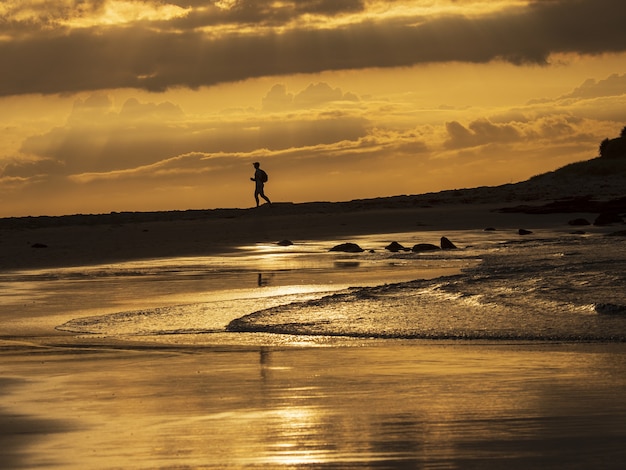 Silhouette of a male running on the rocky shore of the sea under the golden sunset sky