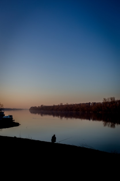 Free Photo silhouette of a male sitting near the water under a clear blue sky in a vertical shot