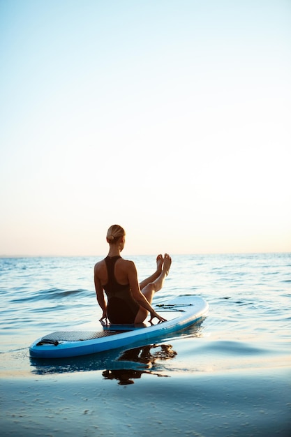 Silhouette of young beautiful girl practicing yoga on surfboard in sea at sunrise.