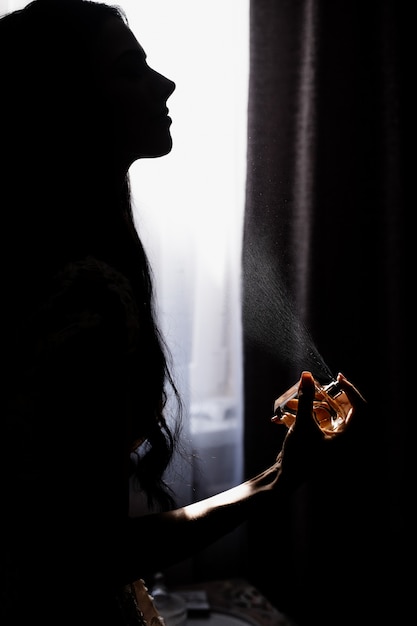 Silhouette of young girl who is perfuming in front of window in darkness