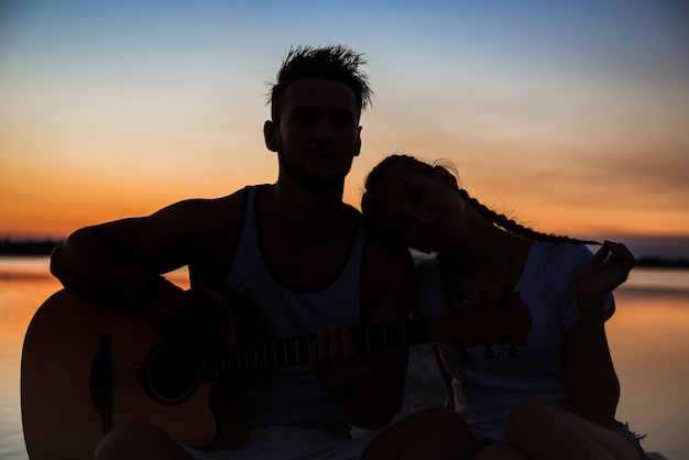 Silhouettes of young beautiful couple resting rejoicing at sunrise near lake