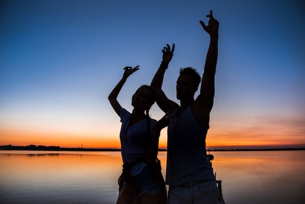 Free Photo silhouettes of young beautiful couple resting rejoicing at sunrise near lake