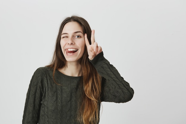 Silly happy woman with long hair do peace sign, winking and show tongue, smiling cheerful