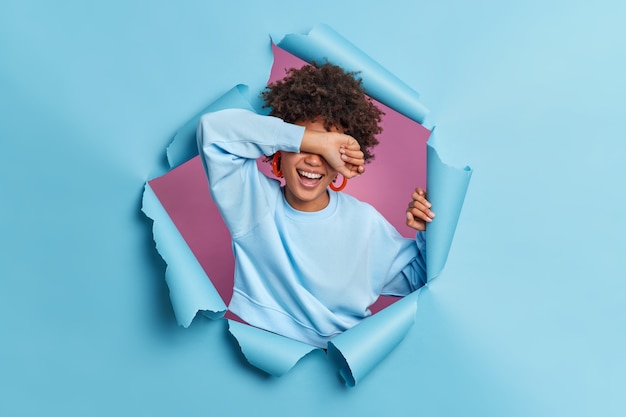 Free Photo sincere curly haired woman covers eyes with arm giggles positively hides face smiles broadly wears casual jumper poses through blue paper wall