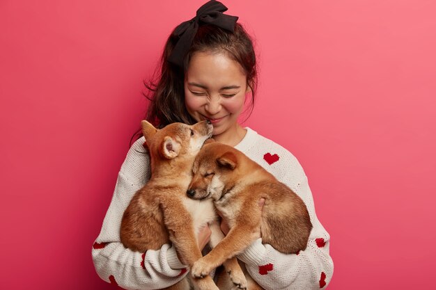 Sincere happy woman plays with two puppies, gets kiss from shiba inu dog, expresses love to animals