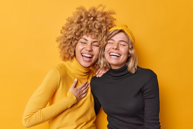 Sincere human emotions concept Positive overjoyed young women have fun laugh gladfully smile toothily cannot stop laughing stand closely to each other dressed casually isolated over yellow wall