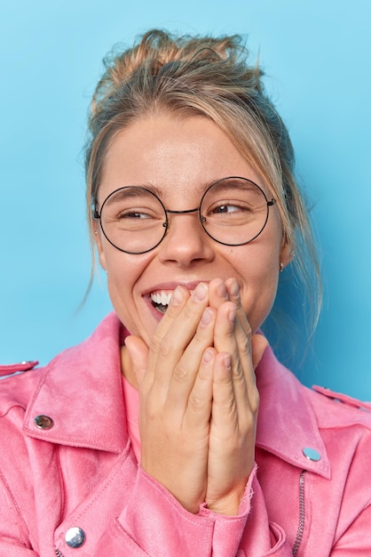 Free photo sincere human emotions. positive young woman covers mouth with hands giggles positively concentrated away feels upbeat wears round spectacles pink jacket poses indoor against blue background