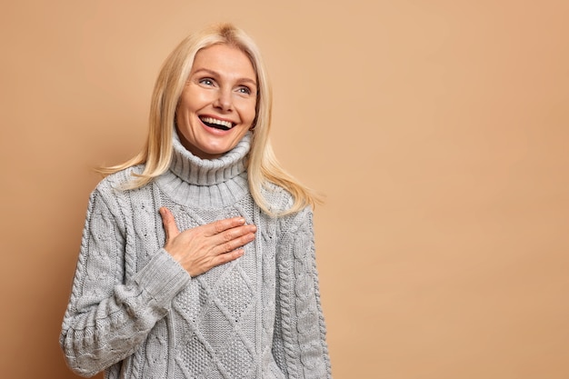 Sincere positive middle aged woman laughs out happily keeps hand on chest smiles broadly has healthy skin minimal makeup reacalls something pleasant wears grey sweater.