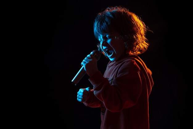 Free photo singing like celebrity, rockstar. caucasian boy's portrait on dark studio background in neon light. beautiful curly model. concept of human emotions, facial expression, sales, ad, music, hobby, dream.