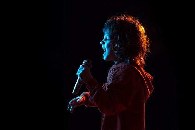 Singing like celebrity, rockstar. Caucasian boy's portrait on dark studio background in neon light. Beautiful curly model.