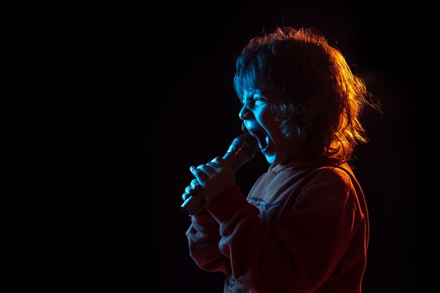 Singing like celebrity, rockstar. Caucasian boy's portrait on dark  wall in neon light. Beautiful curly model. Concept of human emotions, facial expression, sales, ad, music, hobby, dream.