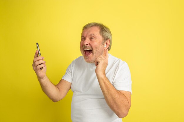 Singing with earphone and smartphone. Caucasian man portrait on yellow studio background. Beautiful male model in white shirt.