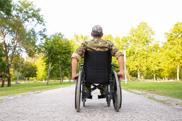 Single disabled retired military man in wheelchair moving down footpath in city park. Back view. Veteran of war or disability concept