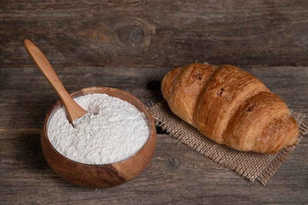 Single fresh plain croissant and bowl of flour wooden surface. High quality photo