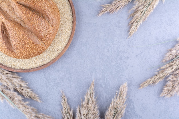 Single loaf of bread in a tray filled with sesame seeds, surrounded with dried feather grass stalks on marble surface