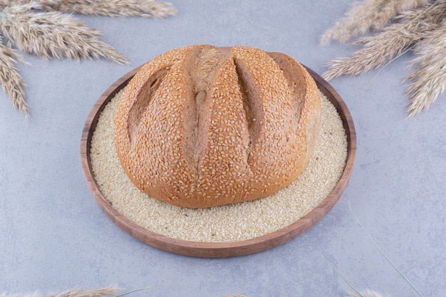 Single loaf of bread in a tray filled with sesame seeds, surrounded with dried feather grass stalks on marble surface