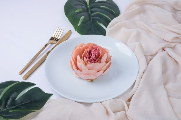 Single rose on white plate with fake leaves and tablecloth.